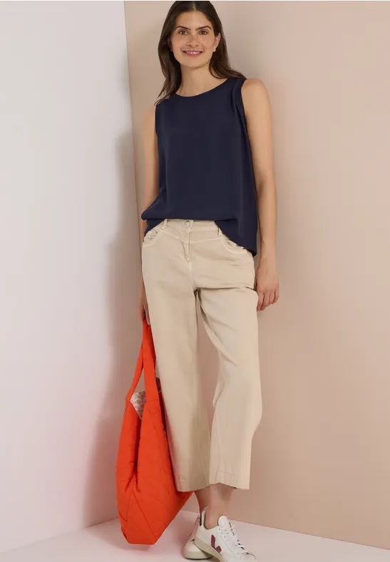 Frau in dunkelblauem Top und weiten, beigen Culotte-Hosen hält eine orange Tasche.