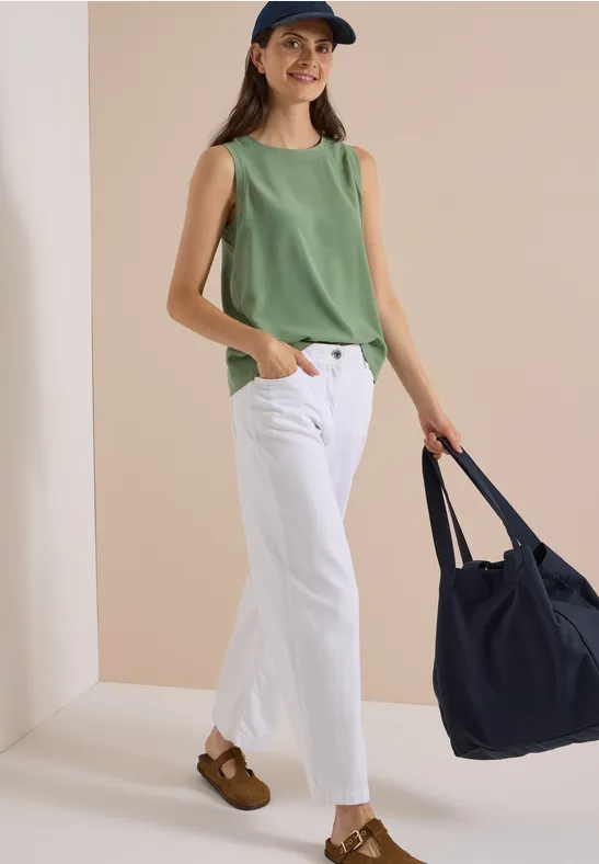 Frau in grünem Top, weißen Hosen, braunen Clogs und Cap hält navy Tasche.