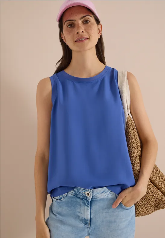 Frau mit pinker Kappe, blauem ärmellosem Top, heller Jeans und Strohtasche vor neutralem Hintergrund.