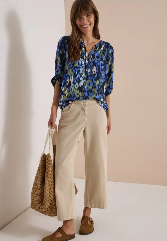 Frau in blau-gemusterter Bluse, beigen weiten 7/8-Hosen, braunen Mokassins und Strohtasche, lächelnd.