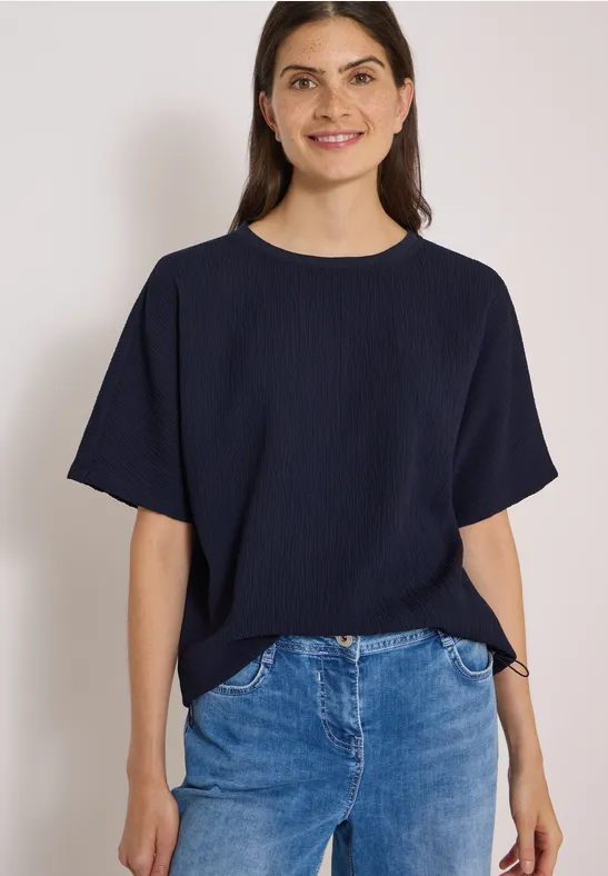 Une femme souriante portant un T-shirt bleu marine et un jean sur fond blanc.