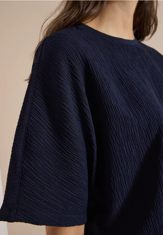 Gros plan d’une personne portant un ample t-shirt bleu foncé à texture plissée sur fond beige.