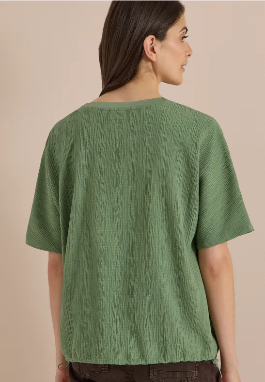 Vrouw van achteren in losse, kreukelige, groene korte mouw top tegen een beige achtergrond.
