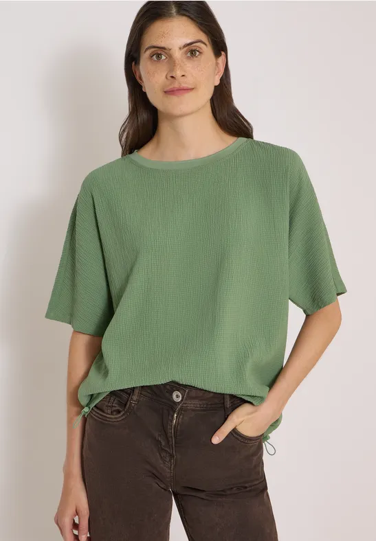Vrouw in groen hemd en donkere broek, hand in zak, met lichte achtergrond.