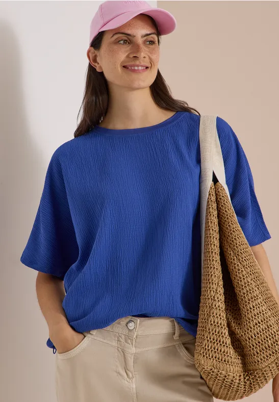 Frau in blauer Bluse, rosa Hut und beiger Hose, lächelt und trägt eine geflochtene Tasche.