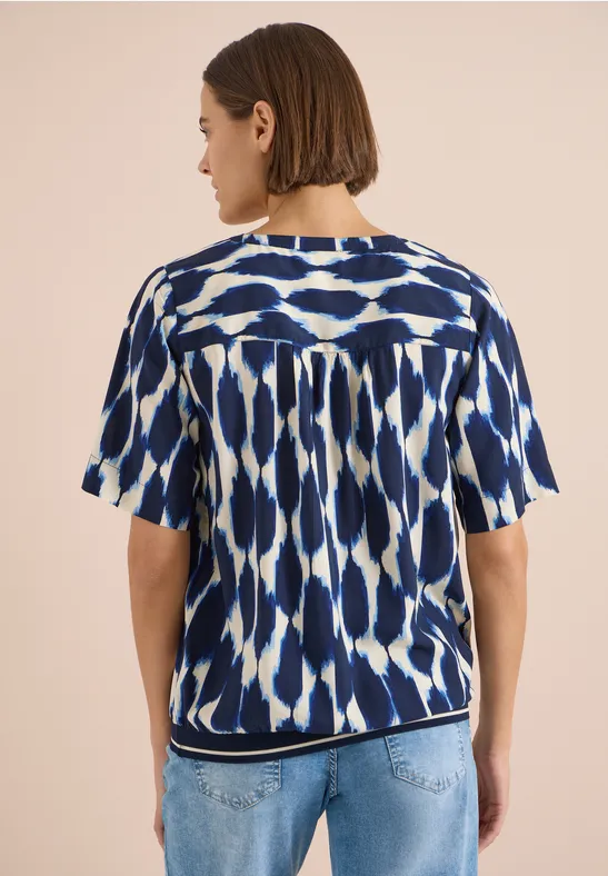 Rückenansicht einer Frau mit kurz geschnittenem Haar, die ein blau-weiß gemustertes Top und hellblaue Jeans trägt.