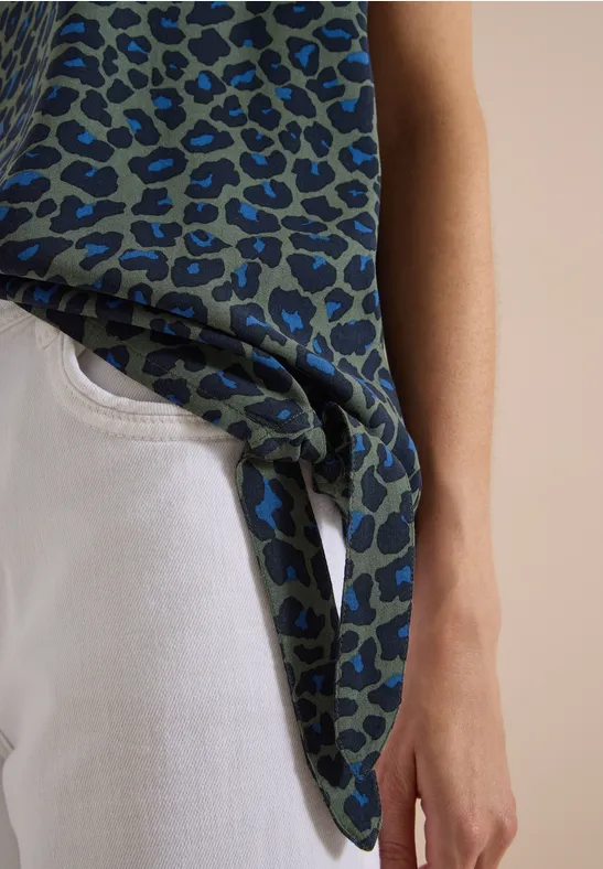 Person trägt weiße Jeans und ein blau-grünes Leopardenoberteil, seitlich locker gebunden.