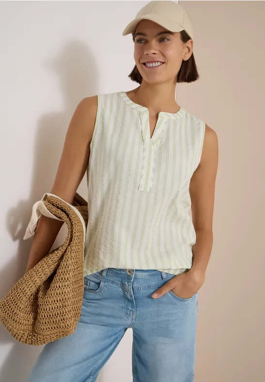 Frau in gestreiftem ärmellosem Top, Jeans und Strohtasche, lächelnd vor heller Wand.