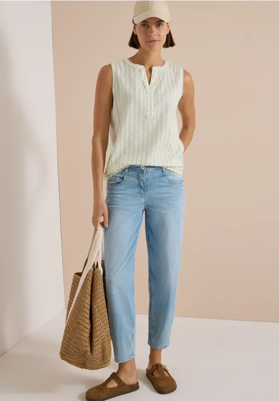 Model trägt beige Cap, weiß-gemustertes Top, hellblaue Jeans, braune Loafers und Korbtasche.