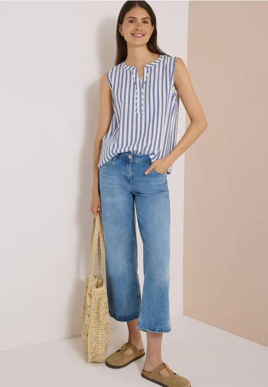 Frau trägt gestreiftes ärmelloses Top, weite Jeans, Basttasche und Slipper.