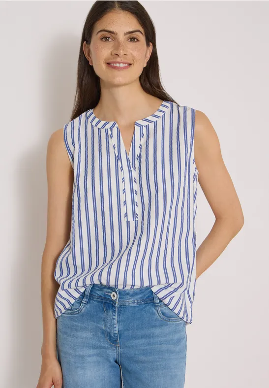 Eine lächelnde Frau in blau-weißer, ärmelloser Bluse mit Knopfleiste, kombiniert mit blauen Jeans.