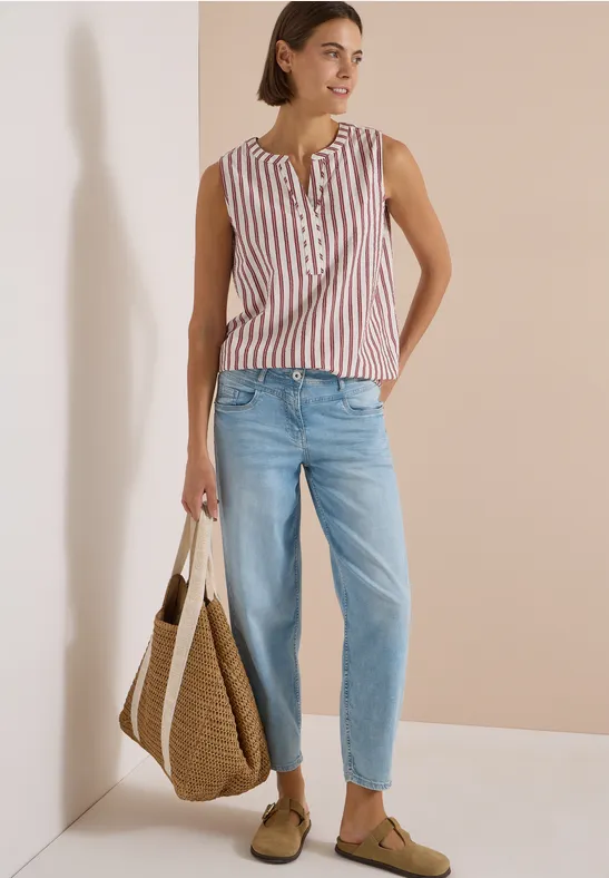 Frau in gestreiftem ärmellosem Top, hellblauen Jeans und Basttasche vor hellem Hintergrund.