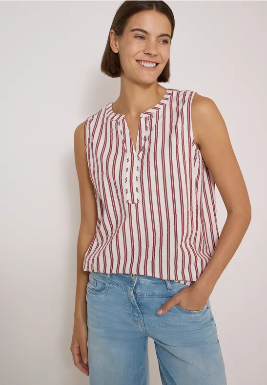 Frau mit kurzem braunem Haar trägt ärmelloses Top mit roten Streifen und blaue Jeans, lächelt vor hellem Hintergrund.