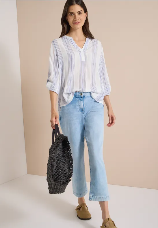 Femme en chemise rayée bleu clair et blanc, jean ample, mocassins beiges, tenant un grand sac noir tissé.