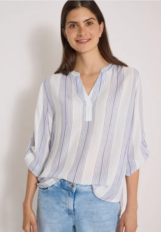 Jeune femme souriante portant un chemisier blanc à rayures bleues et un jean, devant un fond clair.
