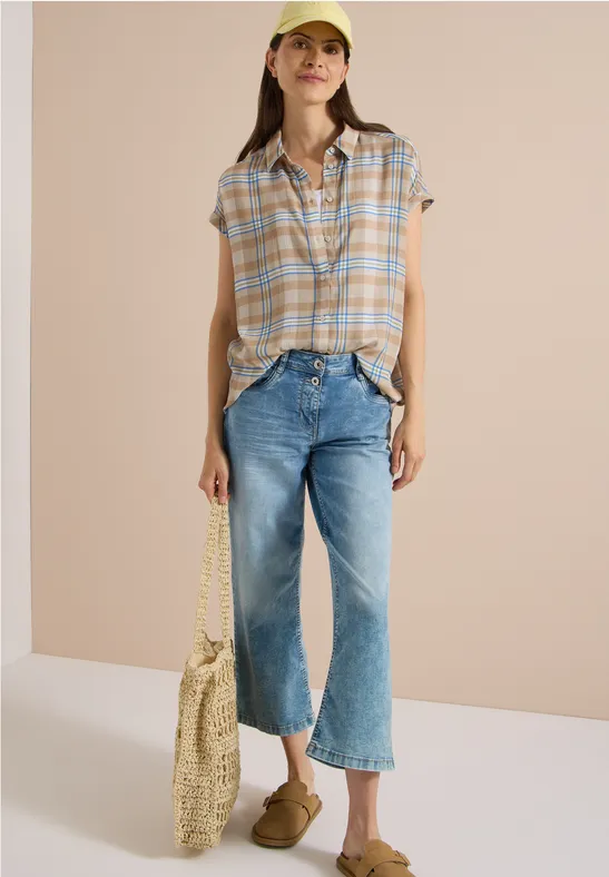 Junge Frau in karierter Bluse, weiter Jeans, Cap, Strohtasche und Pantoletten vor neutralem Hintergrund.