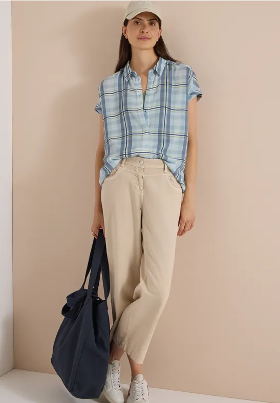 Femme en chemise à carreaux bleue, pantalon en lin beige, baskets blanches, casquette, sac bleu marine.