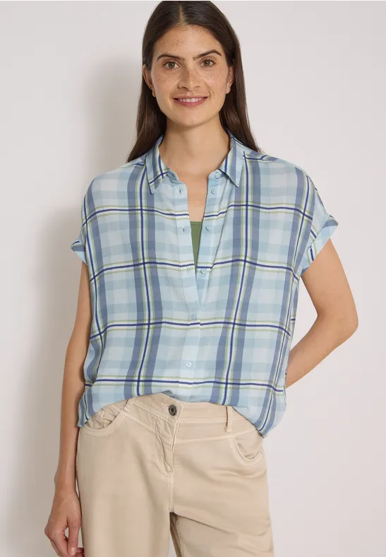Une femme souriante porte une chemise courte à carreaux bleu clair et un pantalon beige, mains dans les poches.