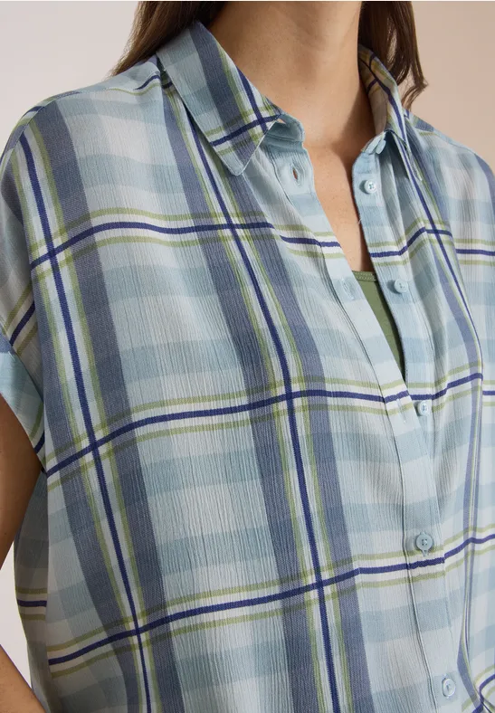 Une personne porte une chemise à carreaux bleu et vert pastel, manches courtes et col ouvert.