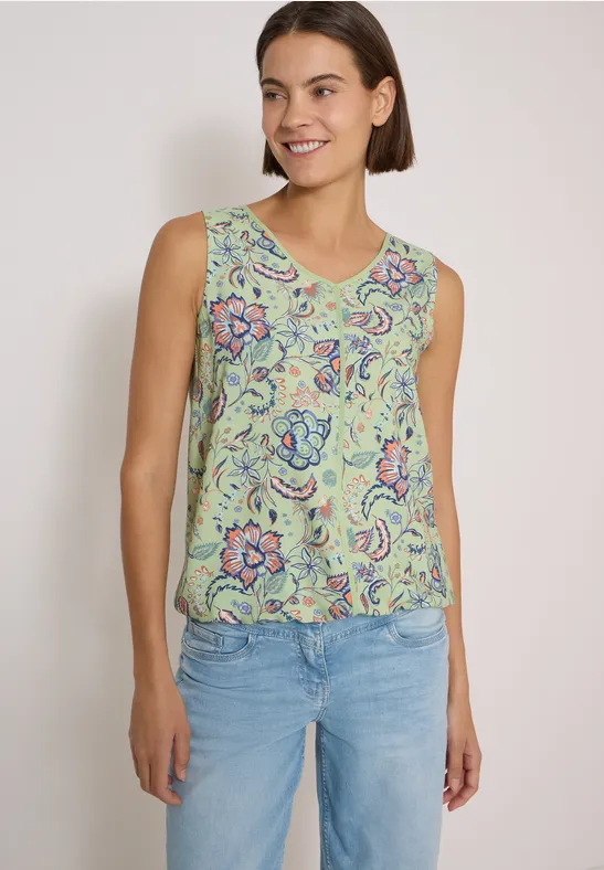 Eine junge Frau trägt ein ärmelloses, grünes Top mit Blumenmuster und helle Jeans vor weißer Wand.