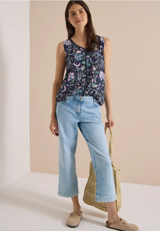 Femme en top sans manches coloré et jean large court bleu clair, tenant un sac en paille.