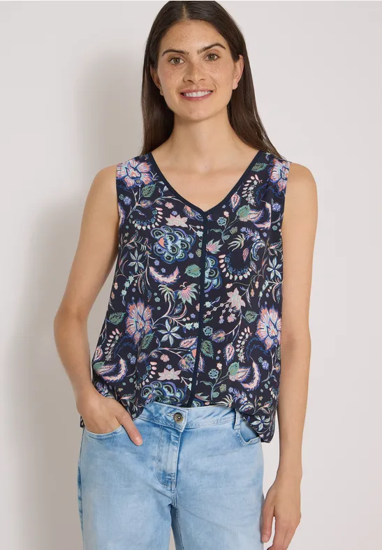 Femme aux cheveux bruns porte un top fleuri sans manches et un jean bleu, sourire naturel.