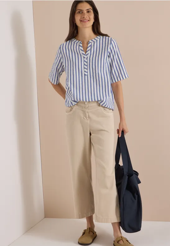 Femme devant un mur neutre, chemisier rayé bleu et blanc, pantalon large beige, chaussures brunes et sac bleu marine.
