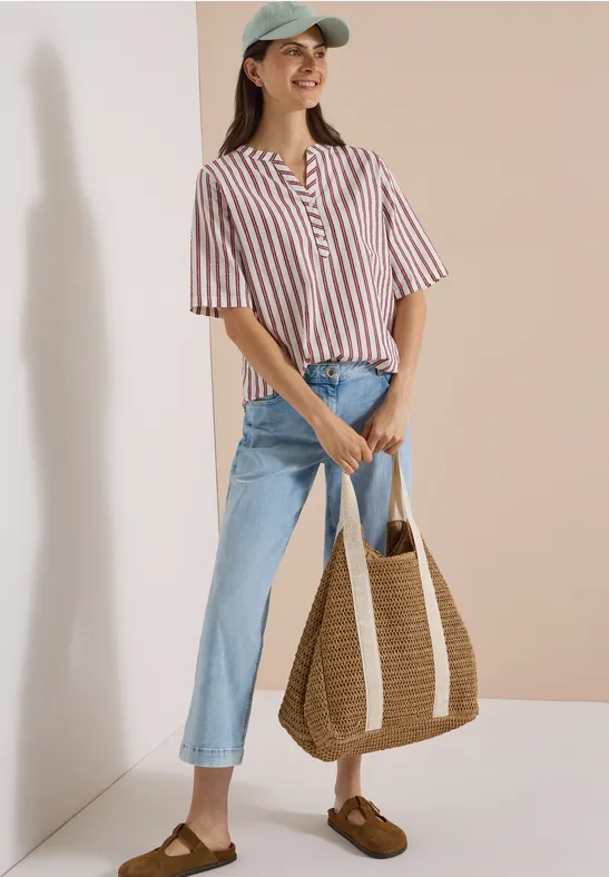Femme en top rayé, jean et casquette verte, tenant un sac en paille, posant devant un fond neutre.