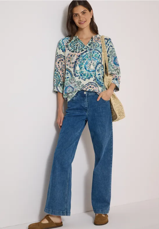 Femme en chemisier à motifs, jeans larges et sac en paille devant fond clair.