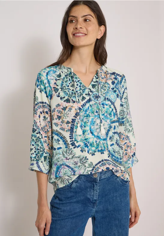 Une femme souriante porte un chemisier coloré à motifs et un jean bleu sur fond neutre.