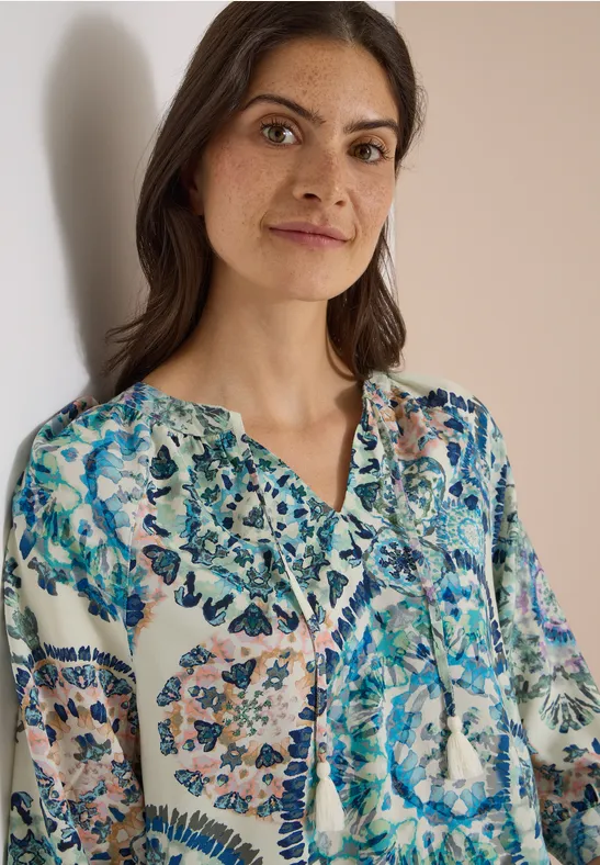 Une femme aux cheveux bruns porte un chemisier blanc orné de motifs floraux bleu-verts colorés.