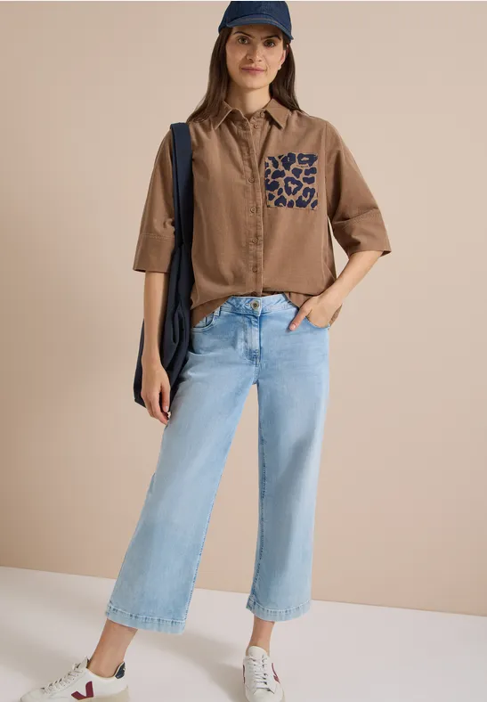 Frau mit Cap, braunem Hemd mit Leoparden-Tasche, hellblauen Cropped-Jeans, weißen Sneakern und Schultertasche vor neutralem Hintergrund.