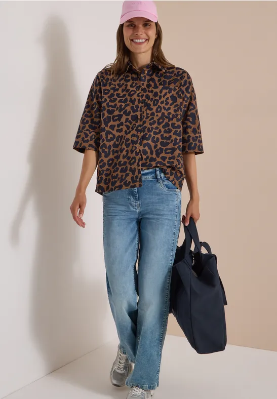 Une femme souriante en casquette rose, chemise léopard, jean, baskets, tenant un tote bag bleu sur fond clair.