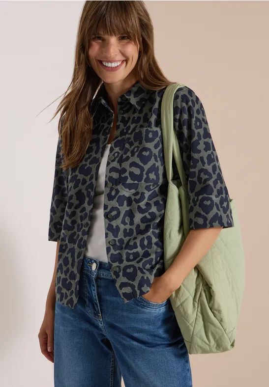 Eine lächelnde Frau trägt eine gemusterte Jacke, weißes Shirt, blaue Jeans und grüne Tasche.