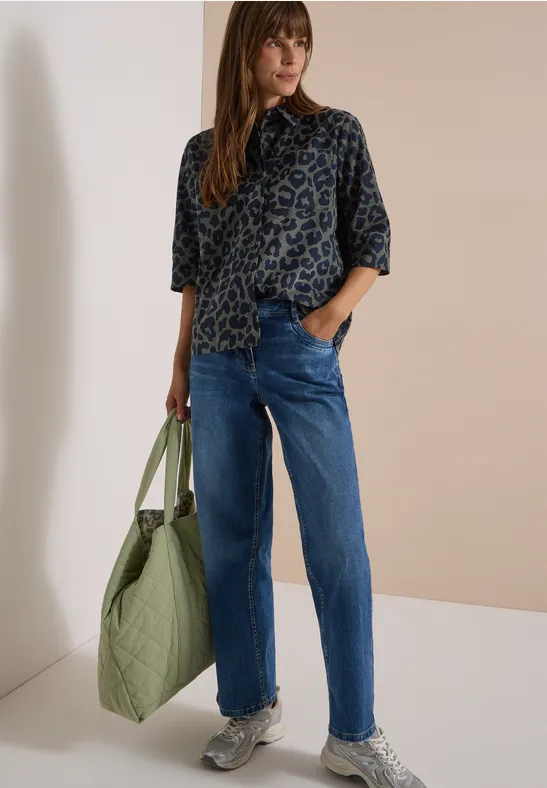 Frau trägt Leopardenmuster-Bluse, blaue Jeans, Sneaker, hält große grüne Tasche