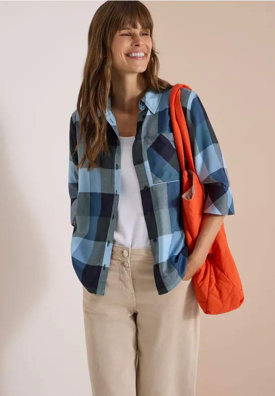 Lächelnde Frau in blau-kariertem Hemd, weißem T-Shirt, beiger Hose und orangefarbener Tasche.
