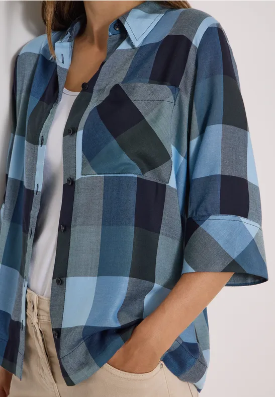 Frau trägt blau-weiß kariertes Hemd mit 3/4-Ärmeln, weißes Top und beige Hose.