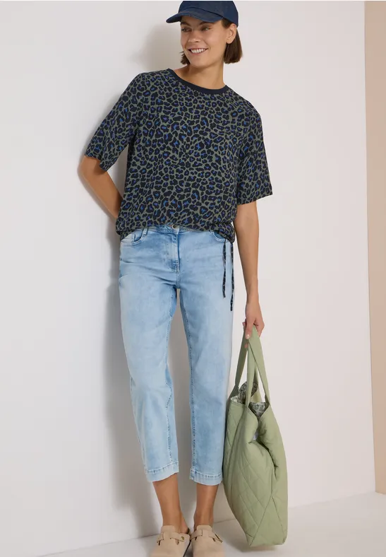 Junge Frau in schwarzem, gemustertem T-Shirt, hellen Jeans, Cap und grüner Stepp-Tasche.