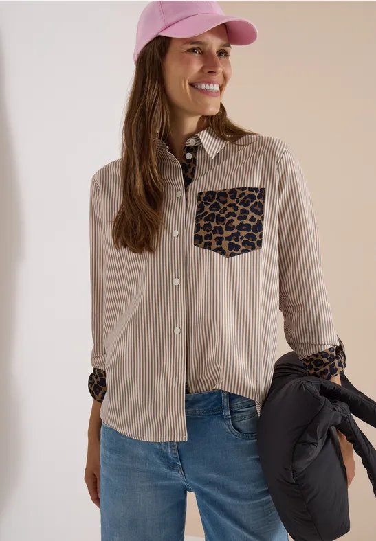 Frau in Jeans, beiger Bluse mit Leopardenmuster-Details, rosa Cap und Pufferjacke in der Hand.