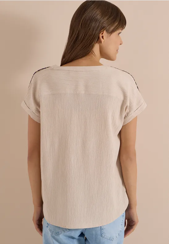 Eine Frau von hinten in einer hellbeigen, kurzärmeligen Bluse und blauen Jeans vor neutralem Hintergrund.
