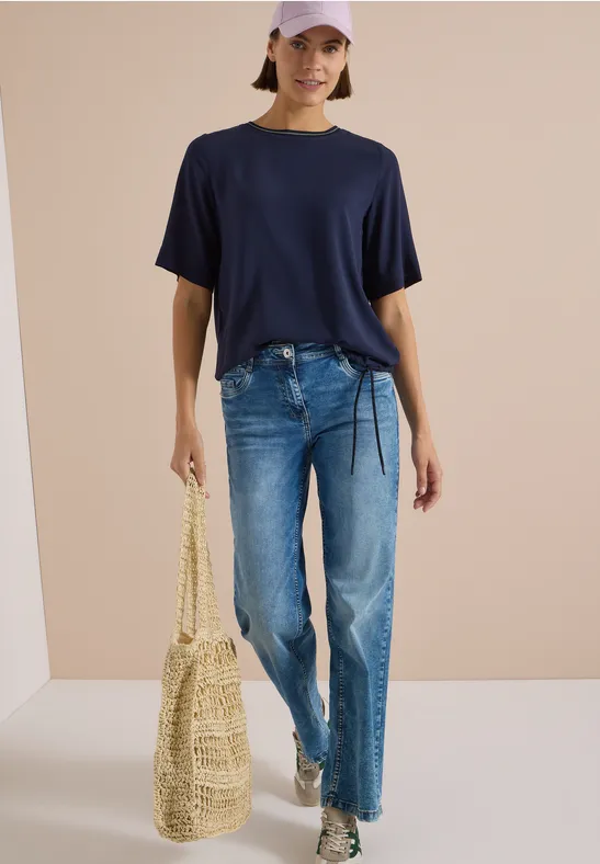Frau in lässigem Outfit: dunkelblaues T-Shirt, Jeans, weiße Sneaker, Strohtasche und rosa Cap.