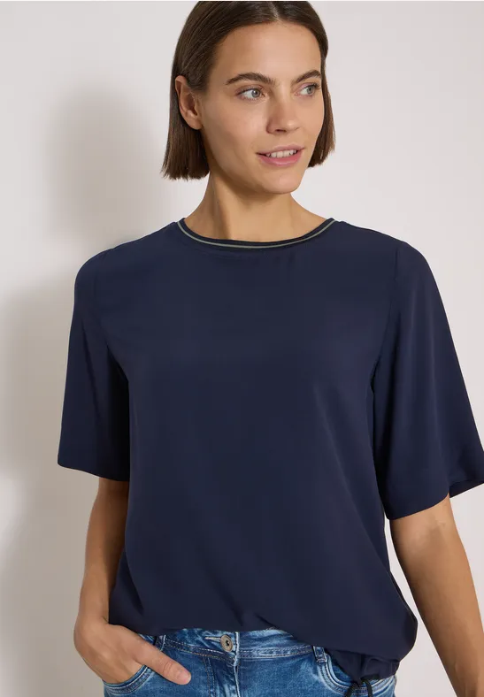 Frau mit kurzem braunen Haar in dunkelblauem T-Shirt und Jeans, schaut leicht lächelnd zur Seite.