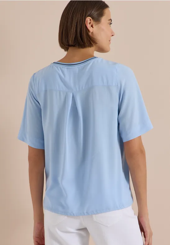 Frau von hinten in hellblauem T-Shirt mit kurzen Ärmeln und weißer Hose vor beigem Hintergrund.