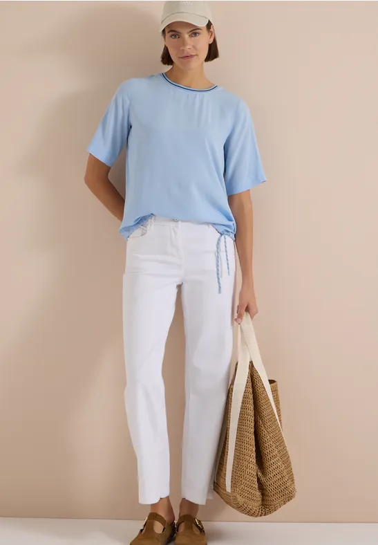 Frau in hellblauem T-Shirt, weißer Hose, Strohtasche und Cap vor beigem Hintergrund.