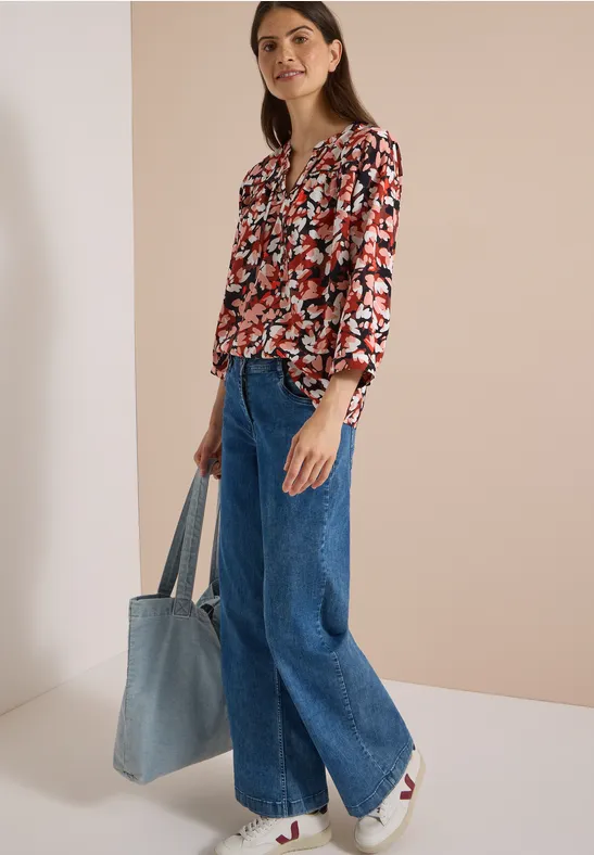 Frau in floraler Bluse und weitem Jeans, weiße Sneaker, hält große Denim-Tasche vor hellem Hintergrund.