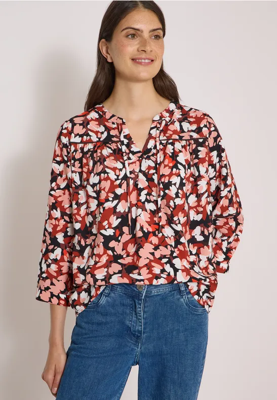Frau trägt Bluse mit rot-schwarzem Blumenmuster und blauen Jeans.