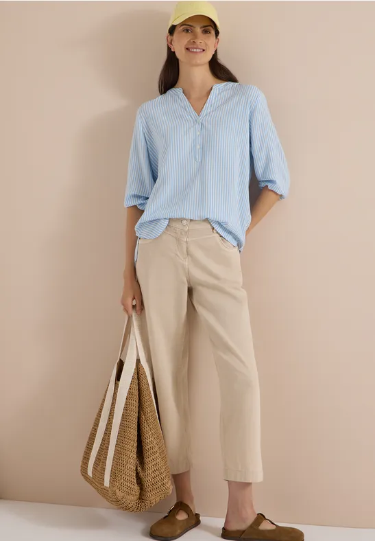 Frau in lässigem Sommeroutfit: hellblaue Bluse, beige weite Hose, Sandalen, Strohtasche und gelbe Kappe.