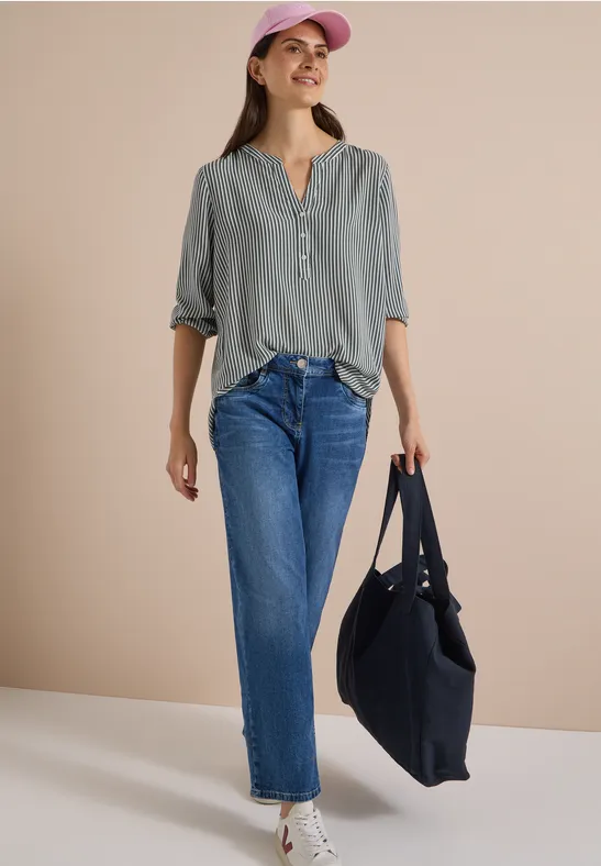 Frau in grauem Oberteil, blauen Jeans, rosa Mütze und schwarzer Tasche vor beigem Hintergrund.