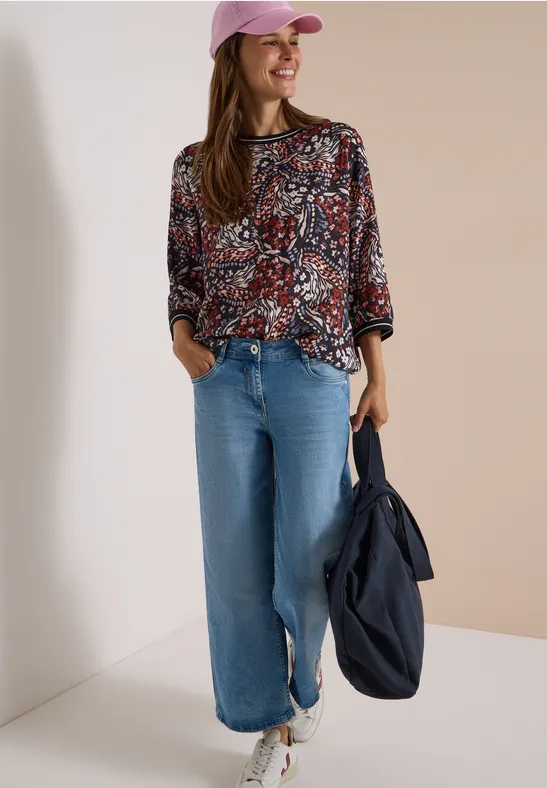 Femme souriante en blouse imprimée, jean large, casquette rose, baskets blanches, sac bleu marine.
