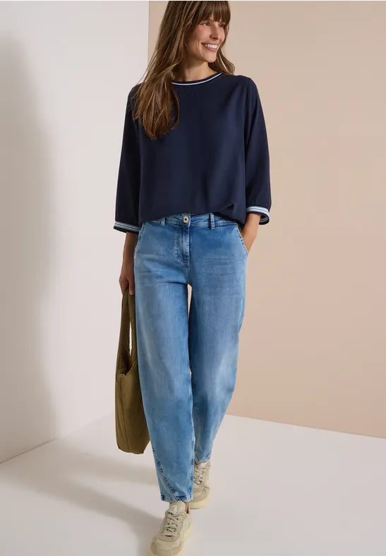 Frau in blauem Pullover und Jeans trägt Tasche und Sneaker vor beigem Hintergrund.